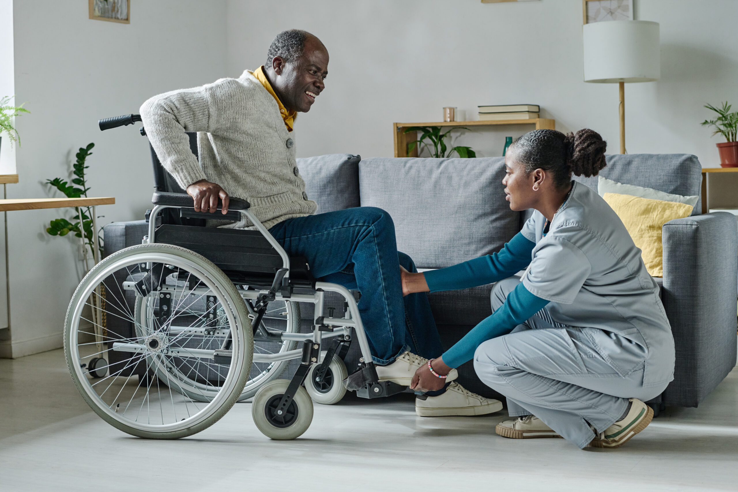 Caregiver helping man to sit on wheelchair African young girl helping senior man with disability to sit on wheelchair, she caring about him at home