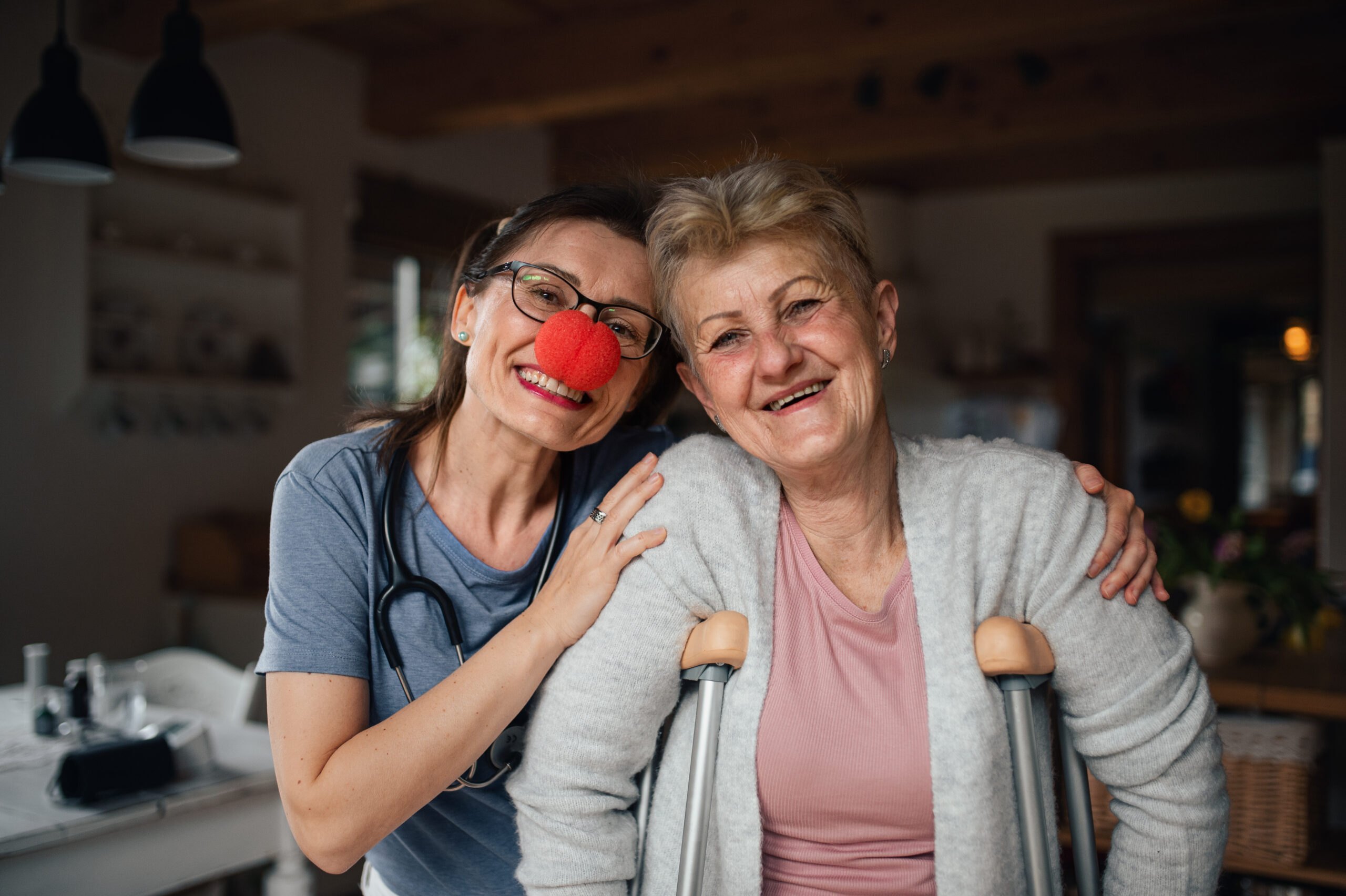 Healthcare worker or caregiver with red nose visiting senior woman indoors at home, looking at camera. A healthcare worker or caregiver with red nose visiting senior woman indoors at home, looking at camera.