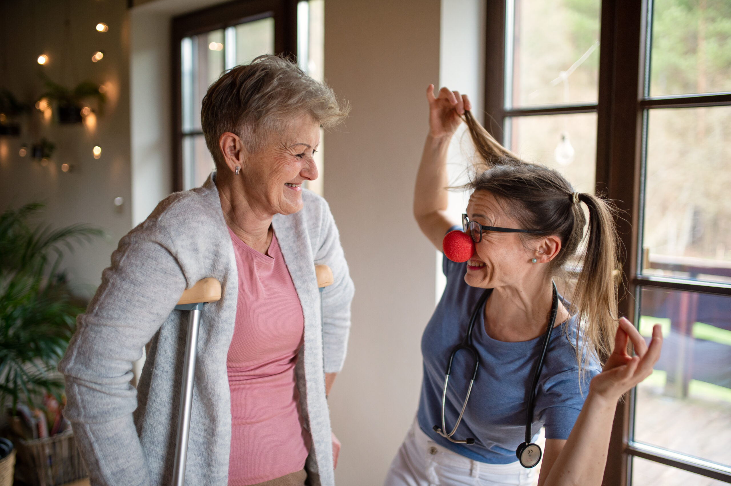 Healthcare worker or caregiver with red nose visiting senior woman indoors at home, having fun. A healthcare worker or caregiver with red nose visiting senior woman indoors at home, having fun.