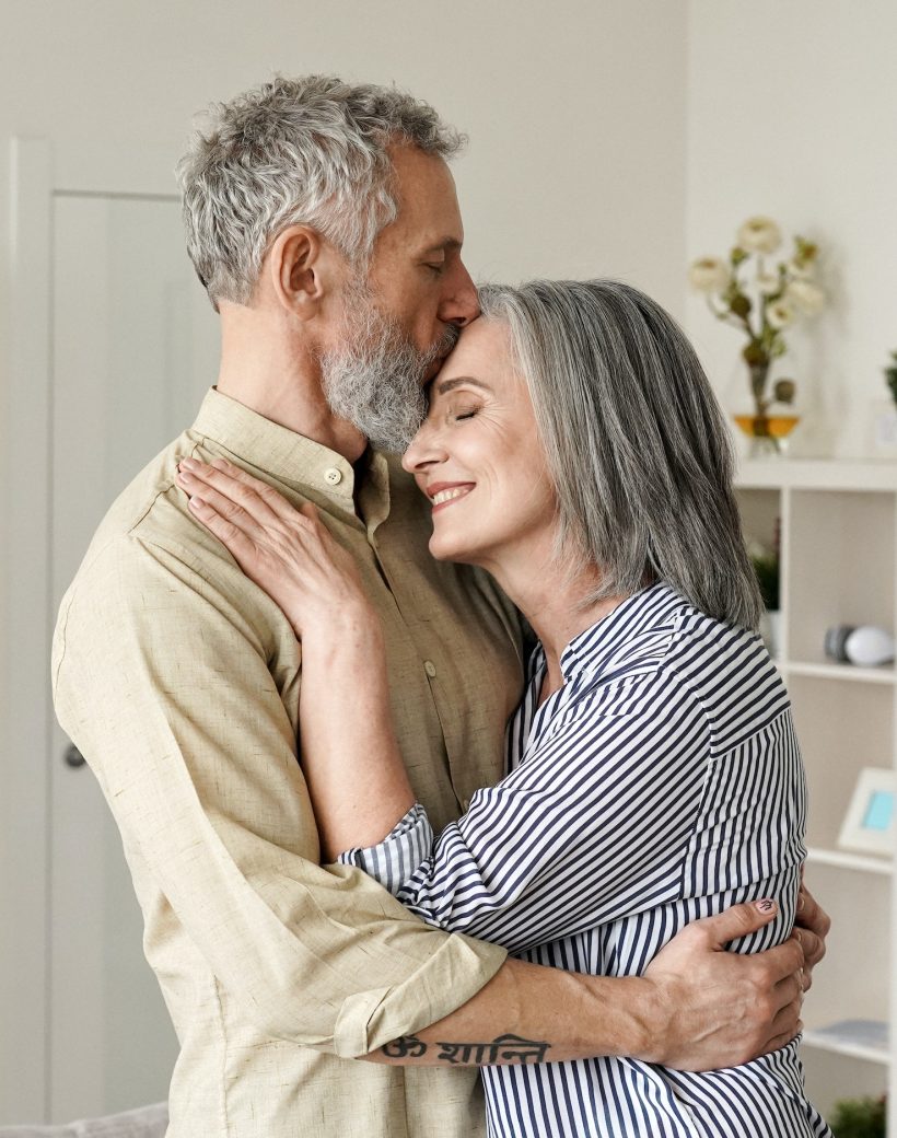 Smiling mid age older couple hugging and kissing standing at home.