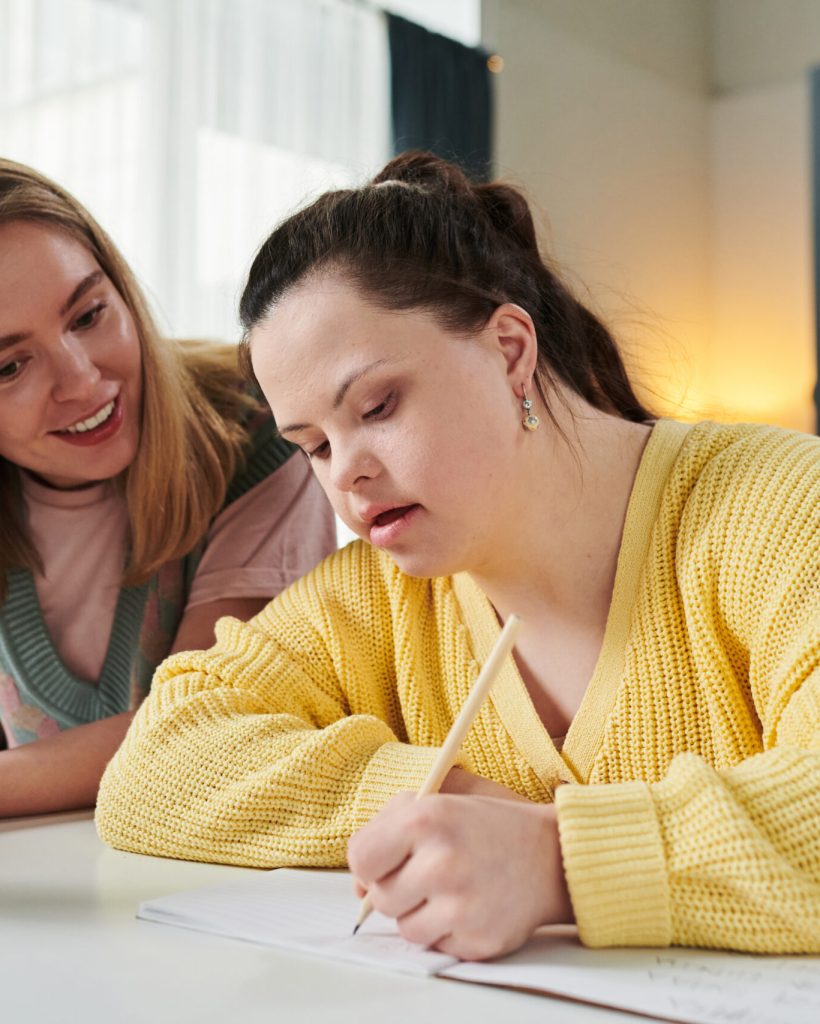 Portrait of young Caucasian woman working as teacher holding textbook giving individual class to girl with Down syndrome